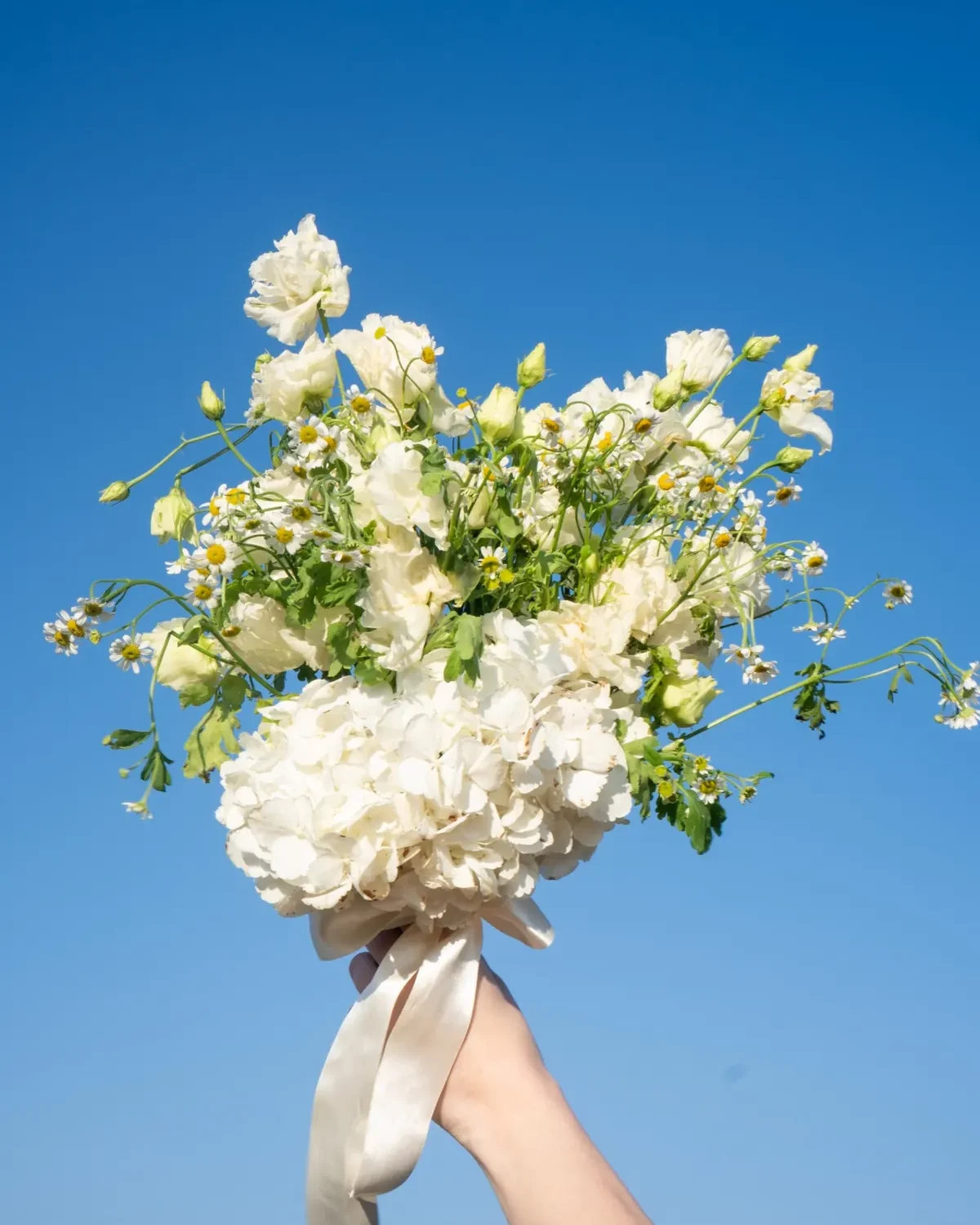 Selene fehér menyasszonyi csokor hortenziával, liziantusszal és kamillával, kézben tartva az ég előtt