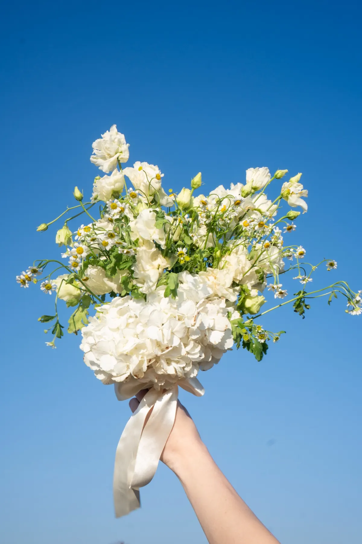 Selene fehér menyasszonyi csokor hortenziával, liziantusszal és kamillával, kézben tartva az ég előtt